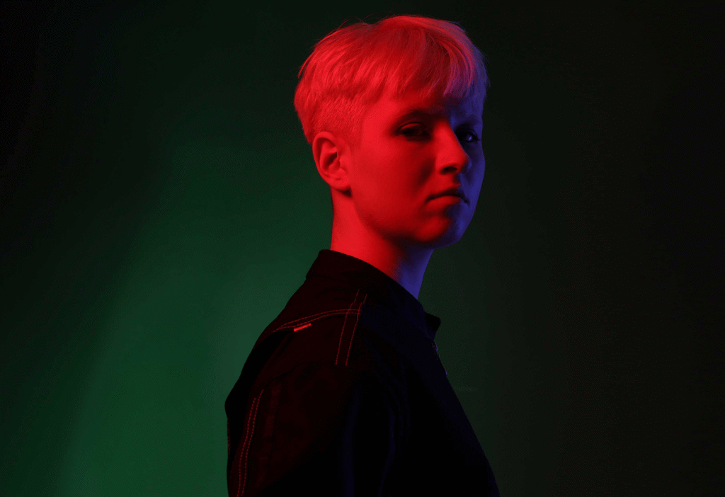 Portrait of a person with short blonde hair lit with dramatic red and blue lighting on a dark green background.