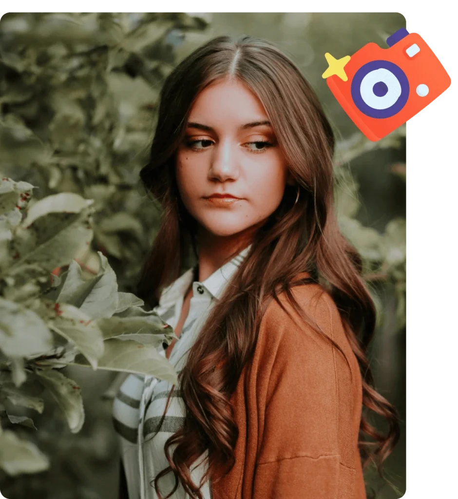 Young woman with long brown hair wearing a striped shirt and brown cardigan, standing among green foliage and looking to the side.