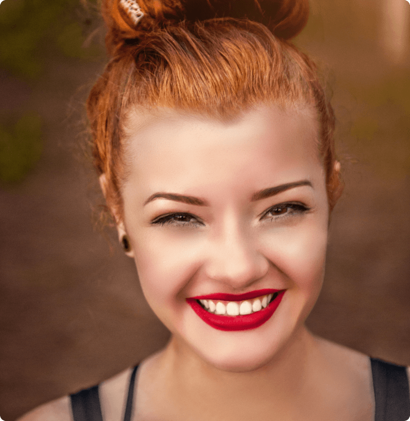 Smiling woman with red hair in a bun, wearing bold red lipstick and dark eyeliner, captured in warm natural light.