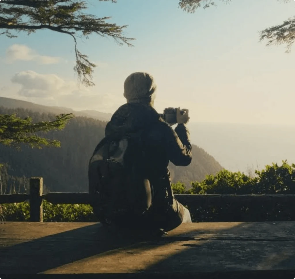 Person with backpack taking a photo of a scenic mountain and ocean view at sunset.