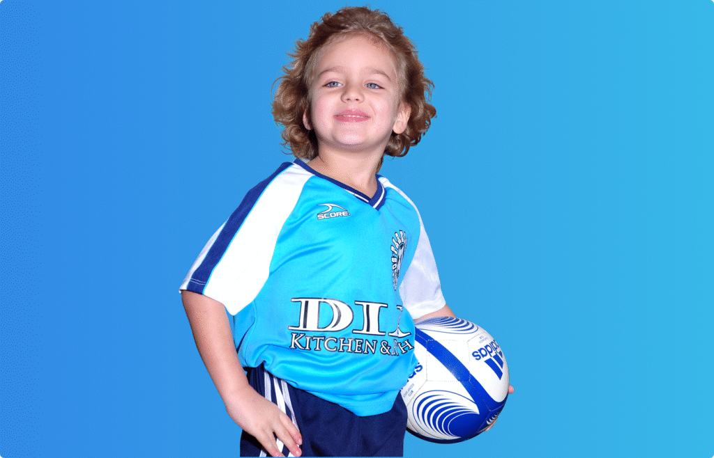 Smiling young child in a blue sports uniform holding a soccer ball against a blue background.